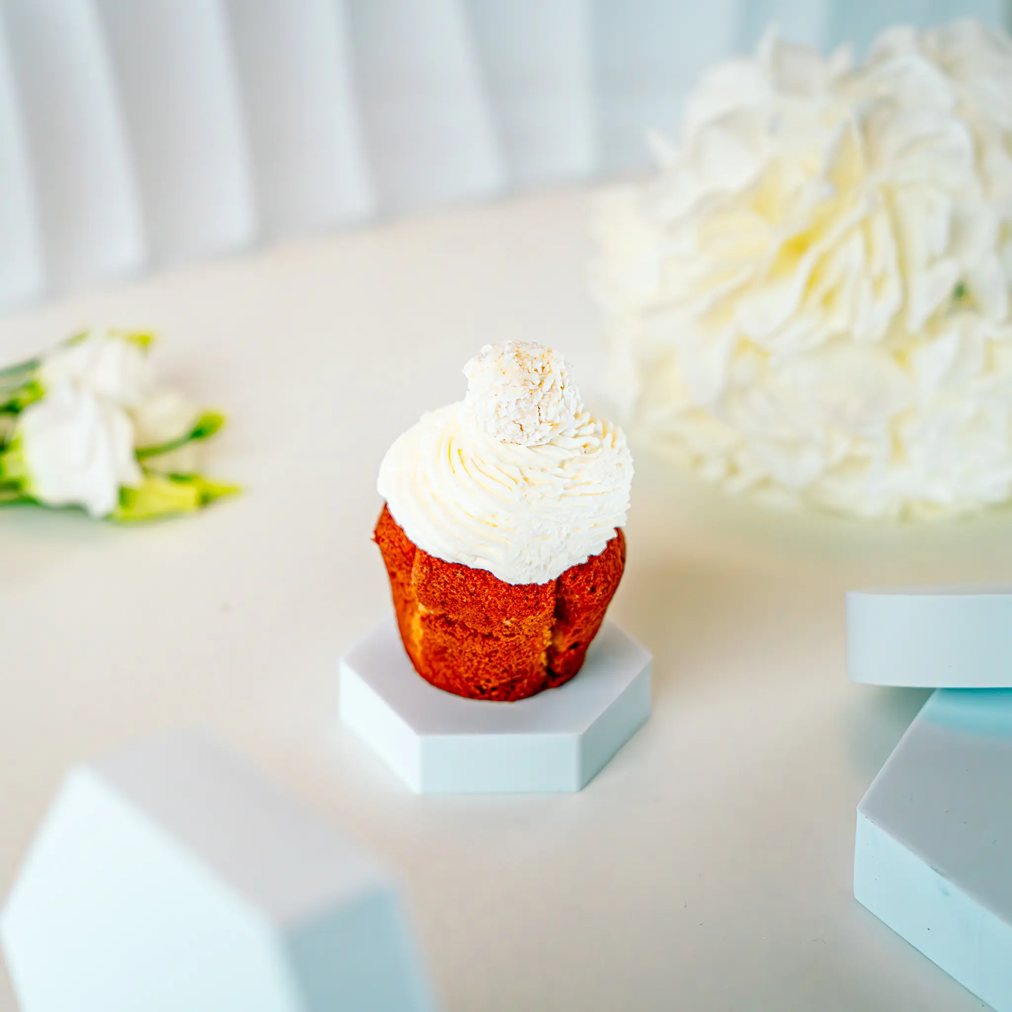 Kokosowy cupcake z białym kremem i pralinką Rafaello na górze, podany na geometrycznej podstawce